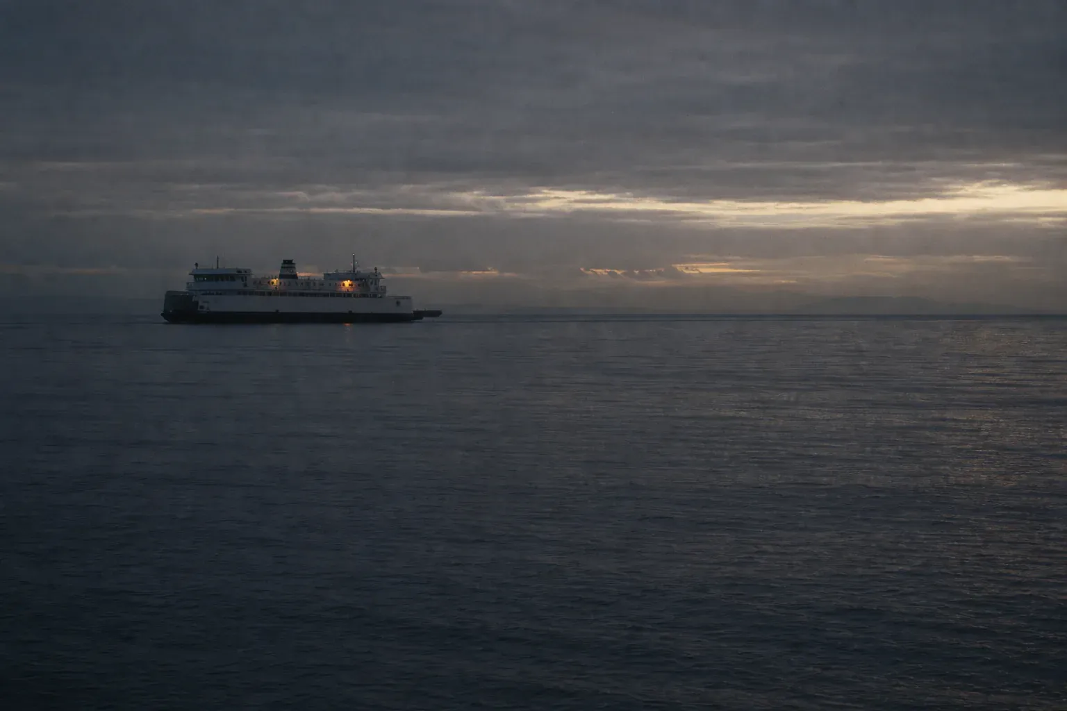 A small white ferry crosses dark water beneath a low bank of cloud before dawn.